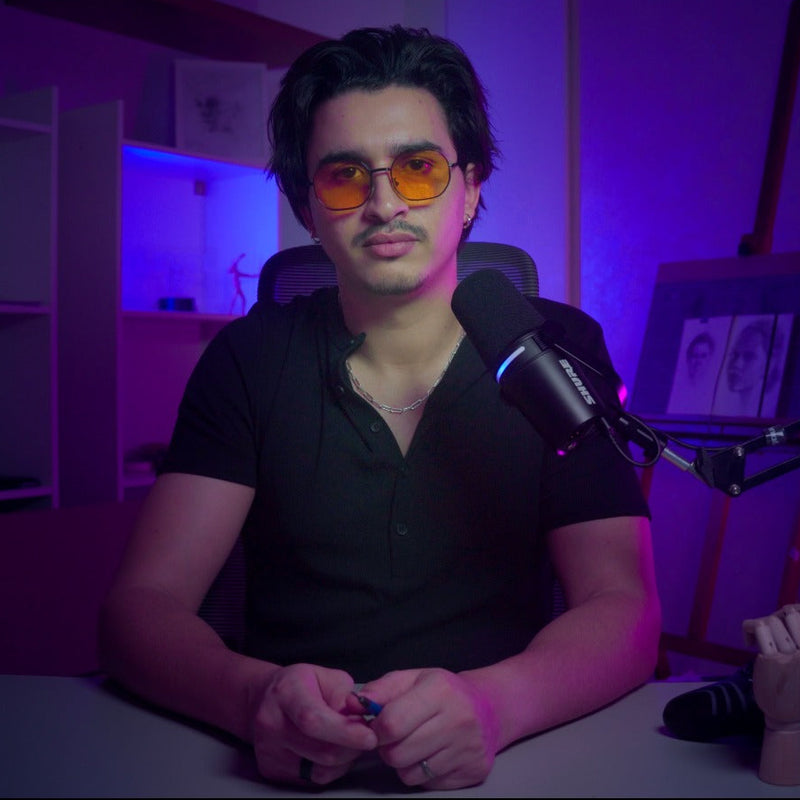 Moe El Boukhari, Founder of Leveling ARtist, wearing orange-tinted sunglasses and a black polo shirt sits at a desk in a dimly lit room with purple ambient lighting, facing a microphone on a boom stand.